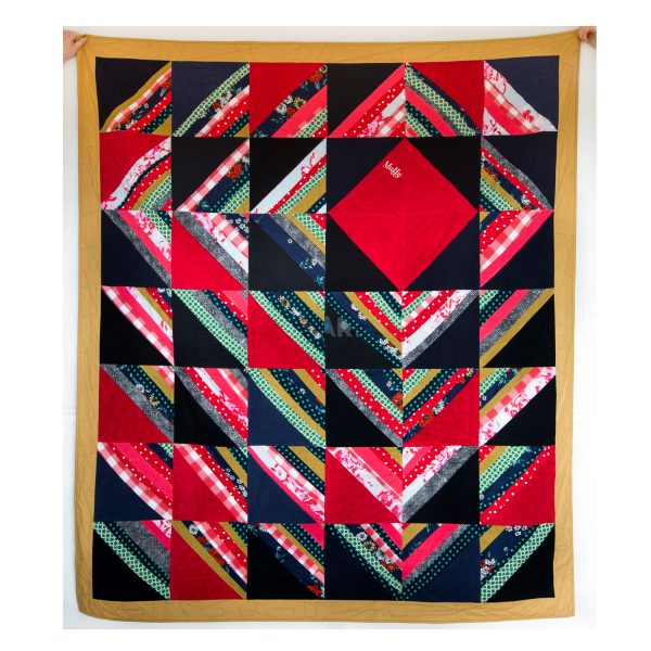 full view of the front of a quilt in reds, greens, blues and blacks. In the upper right, there is a red square made of terry cloth that says 'Muffy'. triangles with strips of color alternating with black triangles radiate outward.