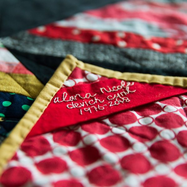 close up of quilt label. White embroidery on a red background, Alana's full name and birth and death dates are hand-stitched.