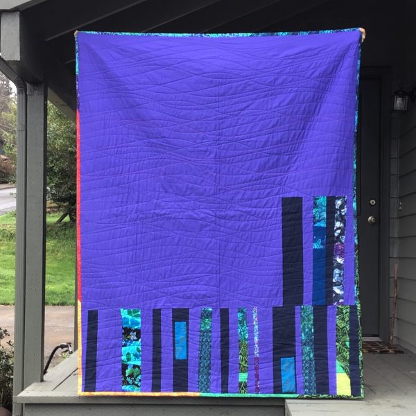 back of quilt. most of the quilt is purple. the bottom third of the quilt features long rectangular strips of fabrics in blues, greens and purples.