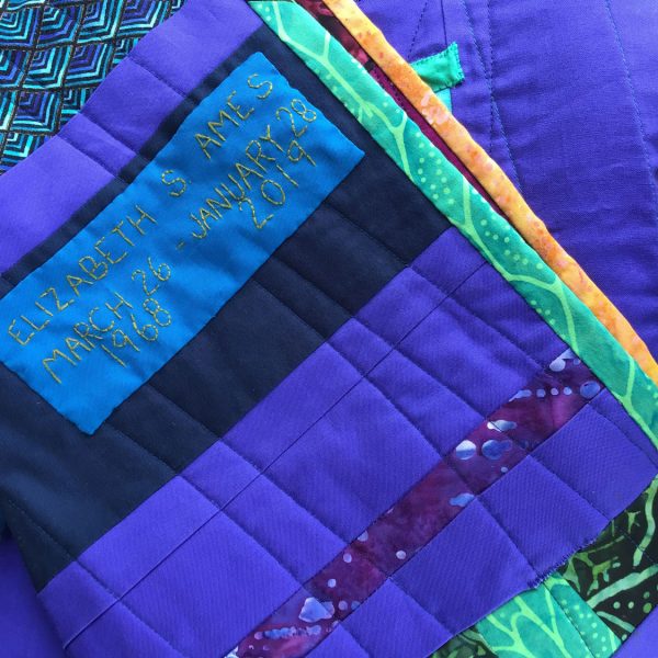 Close up of the back of quilt in blues and purples. A hand embroidered label displays Betsy's full name and birth and death dates.