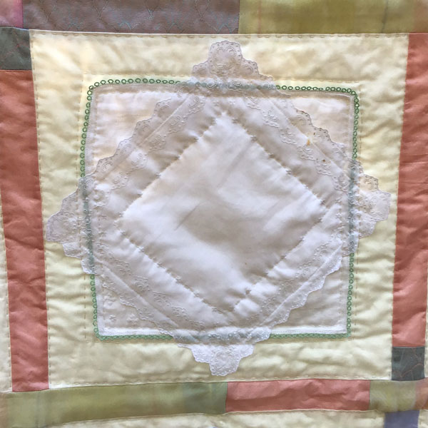 Image is a close-up of one quilt square. Two vintage hankerchiefs overlap on a cream background, bordered by strips of a pastel baby blanket.