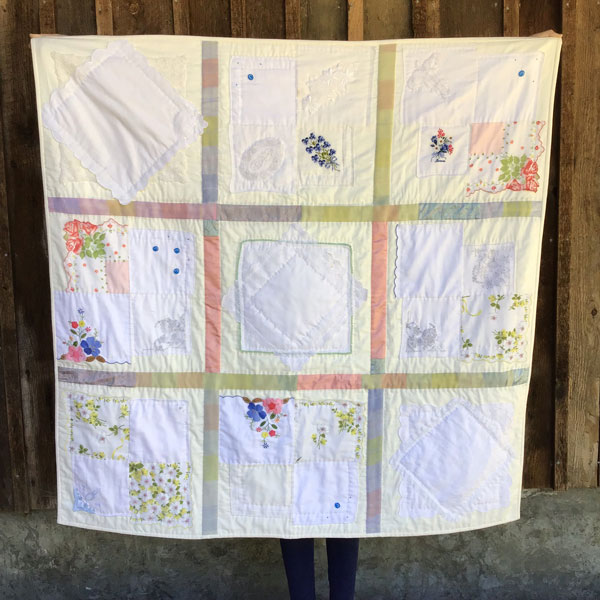 image of a full quilt in chades of cream and pastels. There are 9 blocks sewn from vintage hankerchiefs. Between the blocks are strips of a woven baby blanket in pastel shades of blue, green, pink and yellow.