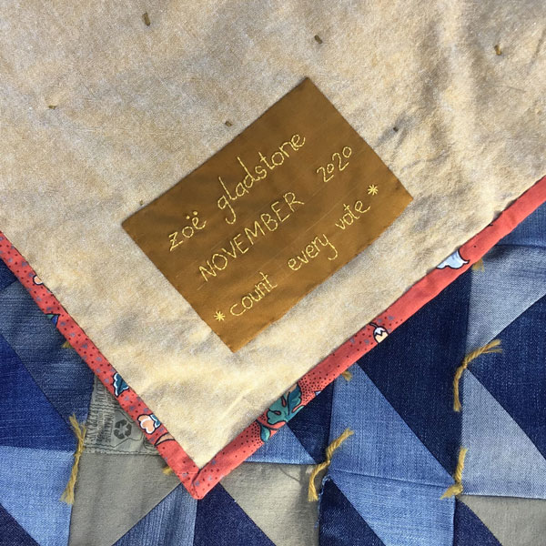 Image is a close up of one corner of the back of the quilt. The backing is made of tan linen, and there is a brown label with yellow stitching. The stitching reads "Zoe Gladstone, November 2020, count every vote'.