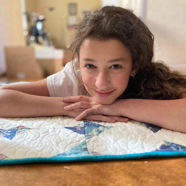 Image shows a tween-age girl with curly brown hair and a sweet smile lying on top of a blue, green and white quilt with her chin on her hands.