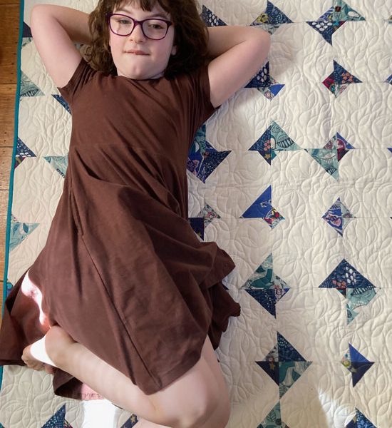 Image shows the quilit on a wooden floor, with a young girl in glasses and a brown dress lying on top of it with her arms behind her head and smiling.