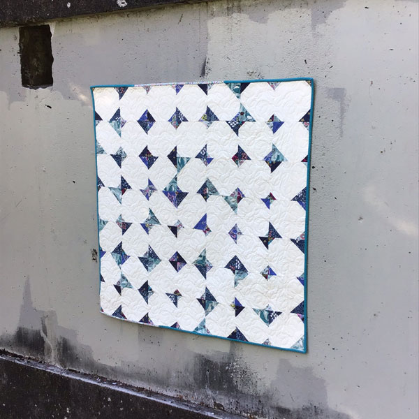 Image shows a full quilt on a concrete wall. Most of the quilt is white, with small triangles of floral fabric in shades of blue and green, and a teal blue binding around the edge.