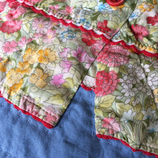 Close up of pink flowered baby dress with red scalloped edges. Dress is on a medium blue quilted background and you can see tiny hand stitching on the dress.