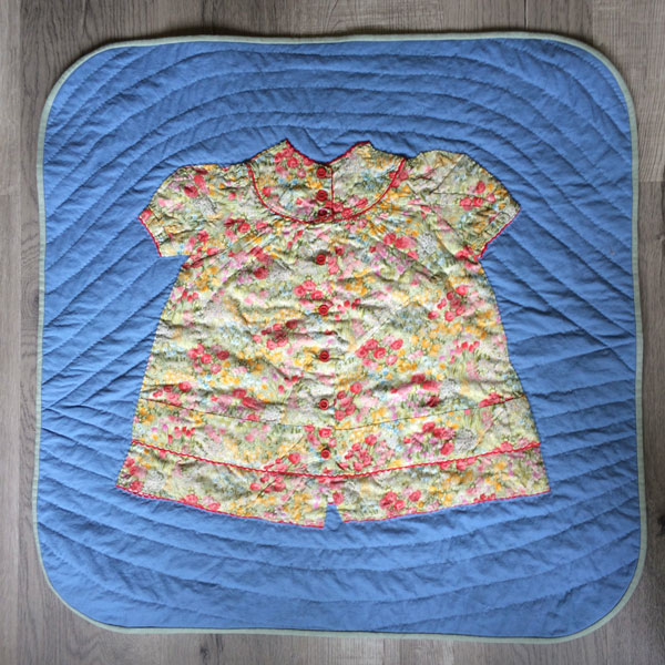 Image shows a small medium blue quilt, slightly larger than the pink flowered baby dress sewn on top of it. Binding is a light sage color.