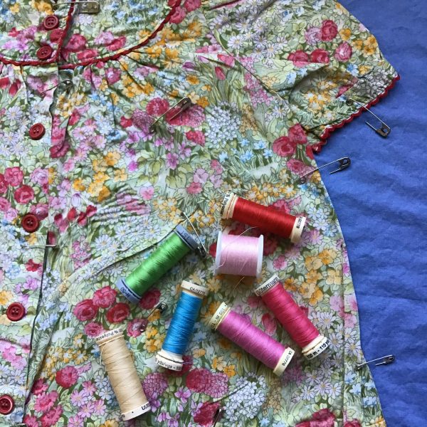 Close up of pink flowered baby dress on a medium blue background. Spools of thread in reds, pinks, greens and blue are are scattered on top of the dress.