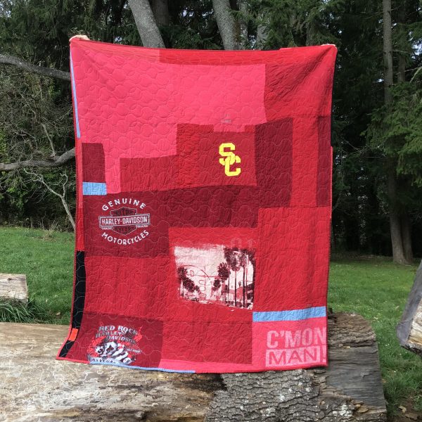 Full view of back of quilt. Design is various shades of red with a few strips of denim blue. Various T shirt graphics are featured with USC and Harley Davidson themes.