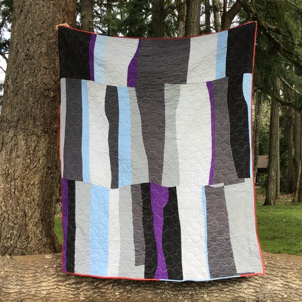 quilt being held up in a forest. The design is organic and thick strips of light and dark grey, purple, and light blue denim.