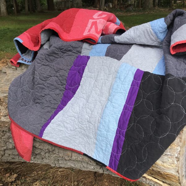 A quilt is piled up in a heap on top of a log. You can see one side of the quilt with strips of grey, purple and blue, and the other side with various shades of red.