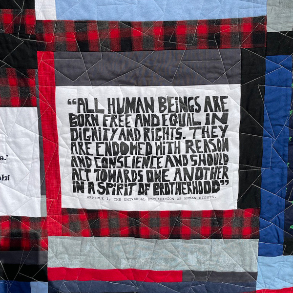 Close up of a red, white, blue and grey quilt square with the words "All human beings are born free and equal in dignity and rights. They are endowed with reason and conscience and should act towards one another in a spirit of brotherhood." Quote is from the Universal Declaration of Human Rights.