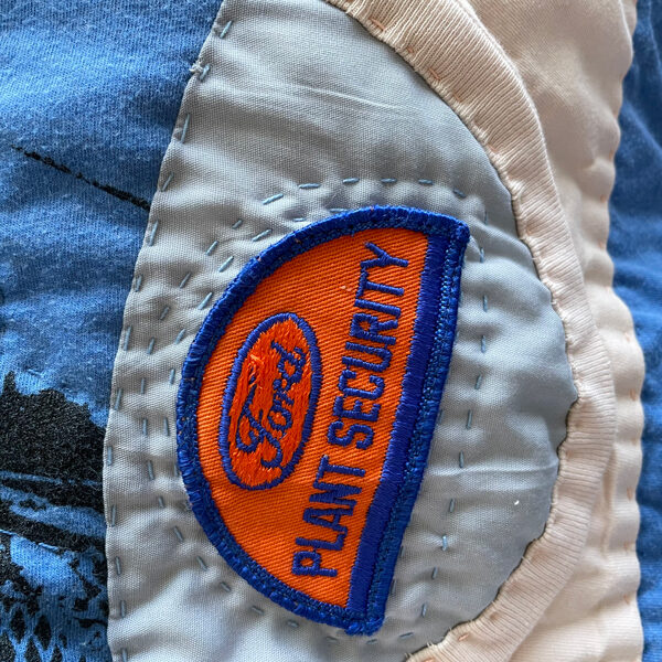 Close up detail of Tshirt quilt, showing hand stitches. Features a half circle blue and orange patch that says "Ford Plant Security".